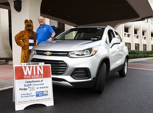 Dr. Craig Hillemeier, 2018 Live United campaign chair, and the Nittany Lion give thumbs up next to the United Way of the Capital Region’s incentive raffle grand prize, a new Chevrolet Trax. Hillemeier is wearing a polo shirt, and the Lion is wearing a striped scarf. In front of the SUV is a sign that says “Win this Vehicle.” They are standing under a portico at the entrance of the Penn State College of Medicine.