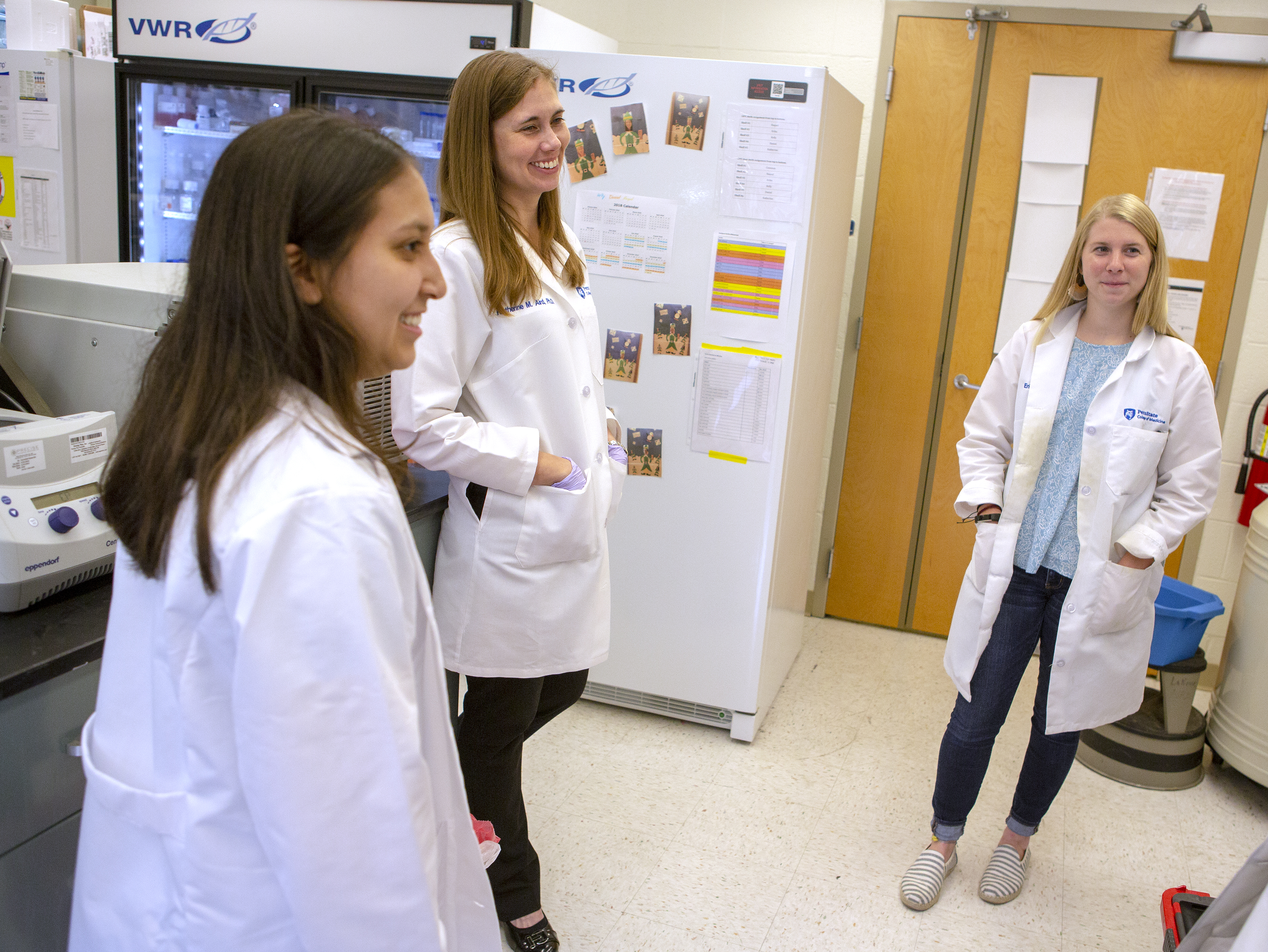 Katherine Aird, center, talks to two members of her research team about their latest work. All three are wearing white lab coats and smiling. Behind them is lab equipment and a refrigerator.