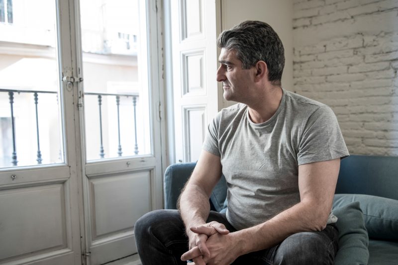 A middle-aged man sits on a couch, looking out the window of the room he’s in.