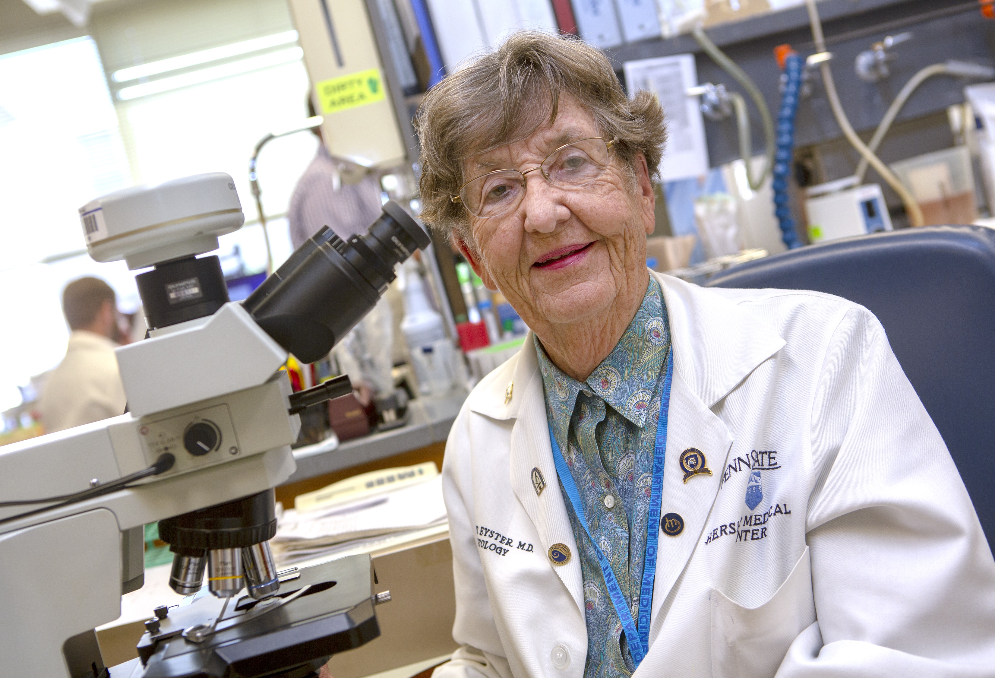 An older woman wearing a white lab with the Hershey Medical Center logo on it sits next to a microscope in a research lab. Behind her is a man at a computer, lab equipment and binders on a shelf.