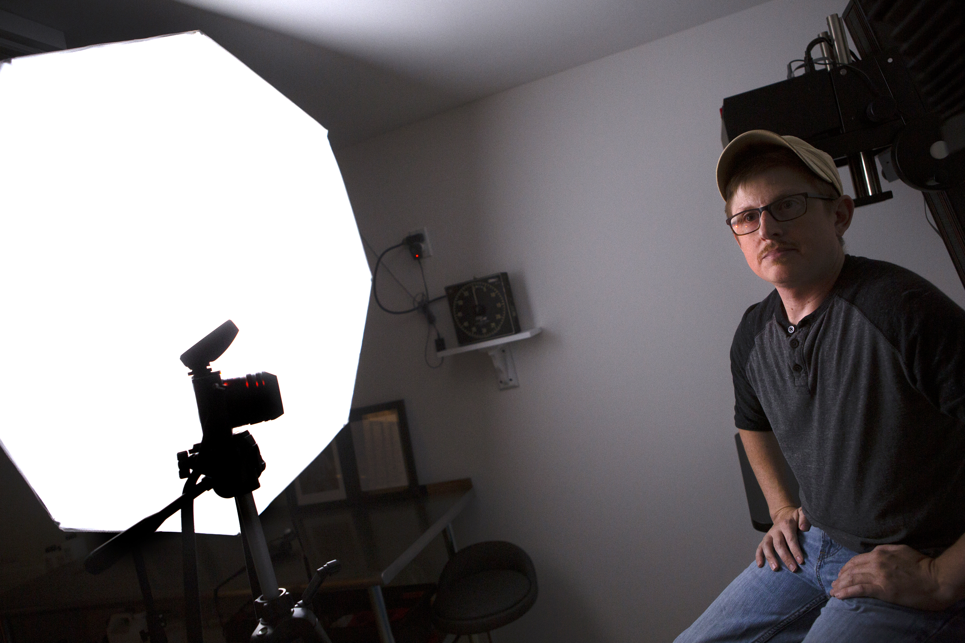 A man wearing a baseball cap, T-shirt and jeans sits in front of a camera on a tripod and an umbrella lamp. In the background is a table, a stool and a timer.
