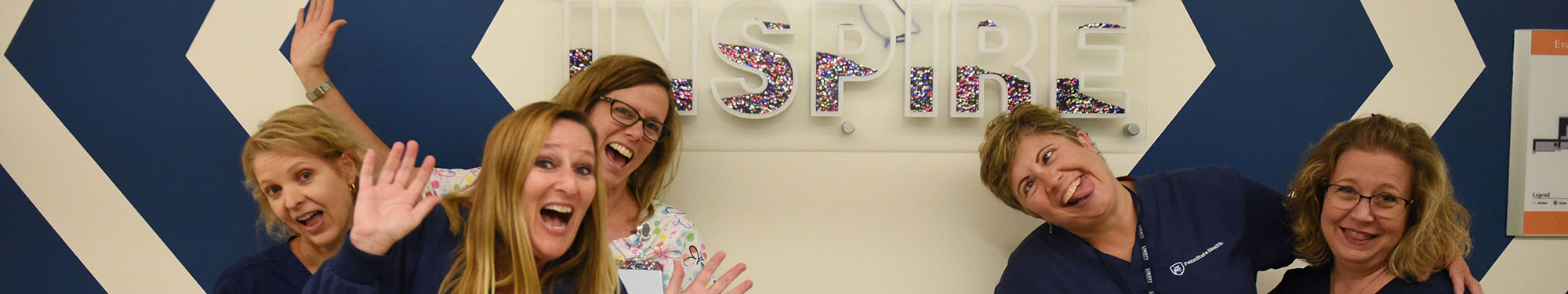 A group of nurses are seen in the Survivorship Clinic at Penn State Children's Hospital. They are making silly faces and gesturing to a large depiction of the word INSPIRE on the wall behind them.