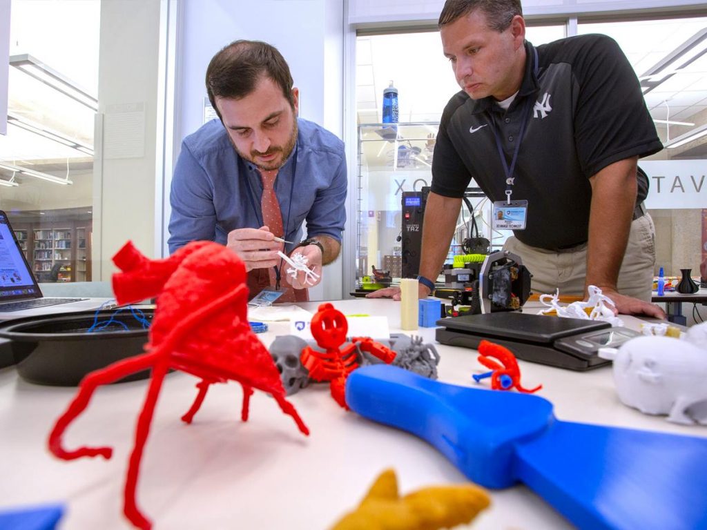 Russell Dicristina, right, looks over at Mike Cote working on a 3D model during the Day of Making event at Harrell Health Sciences Library in August 2018. The two are pictured at a table with brightly colored 3D printed objects in front of them.