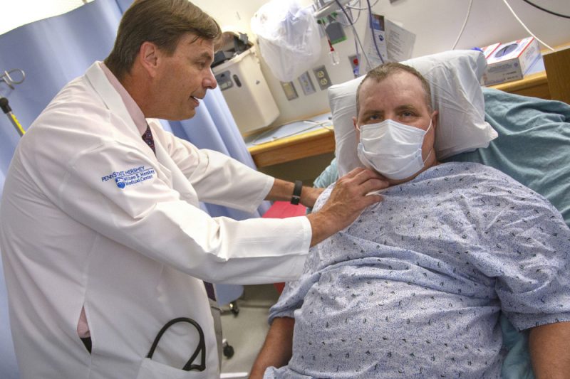 A physician in a white coat stands next to a patient who's in a hospital bed. The doctor's hands are placed on the patient's shoulder and neck area.