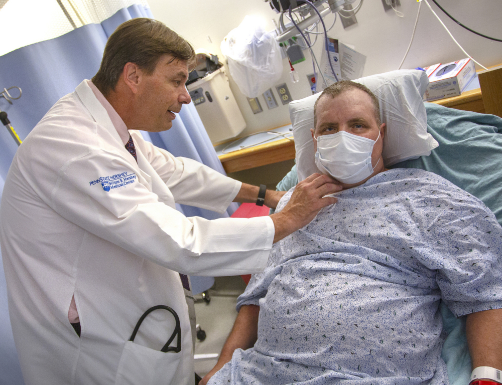 A physician in a white coat stands next to a patient who's in a hospital bed. The doctor's hands are placed on the patient's shoulder and neck area.