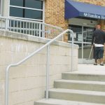 A man with a crutch walks to the door of the Hamilton Health Center. Concrete steps and railings are in the foreground.