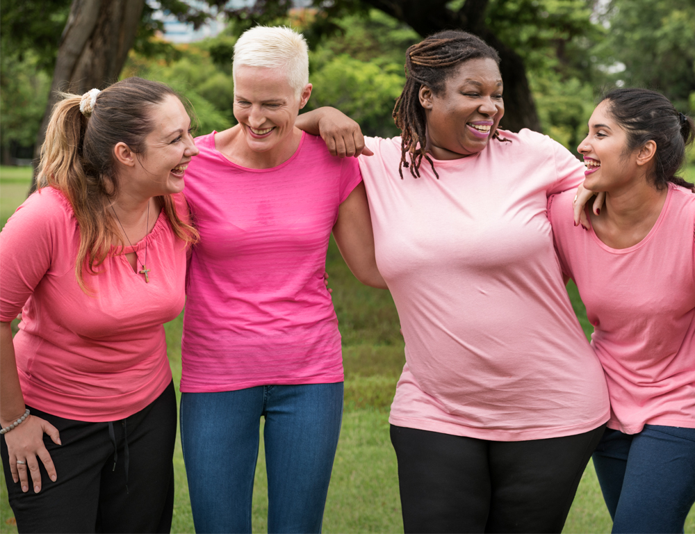 Four women wearing pink t-shirts stand with arms around each other, smiling. Grass and trees are in the background.