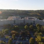 An aerial view of the Milton S. Hershey Medical Center and Penn State College of Medicine campus. The Crescent building surrounded by trees with fall covers and a mountain in the background. Cars are in the parking lot. A sunspot highlights the left side of the sky.