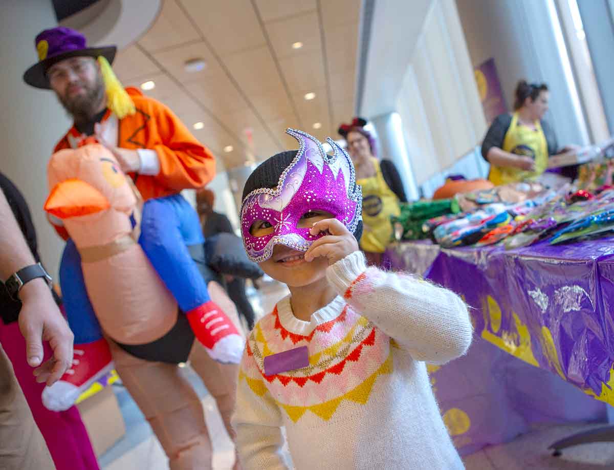 A young girl smiles and holds a mask to her face. She is wearing a sweater and a knit cap. Behind her is a man in a costume as a person riding a turkey and a table of costumes and assorted Halloween items.