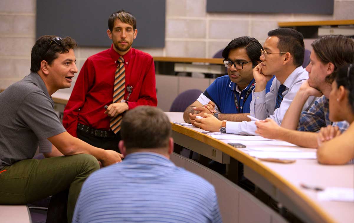 Six medical students sit at a conference room table and listen to a man talk at the American Medical Association’s “Accelerating Change in Medical Education” conference. Behind him Dr. Joshua Davis of Penn College of Medicine, stands and listens.