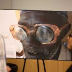 A man and woman look at a large photograph of a man from Ghana on an easel. The man from Ghana is wearing glasses with lenses that are falling out. He has one tooth visible in his mouth. The man looking at the photo is wearing a plaid shirt, while the woman is wearing a black top and glasses.