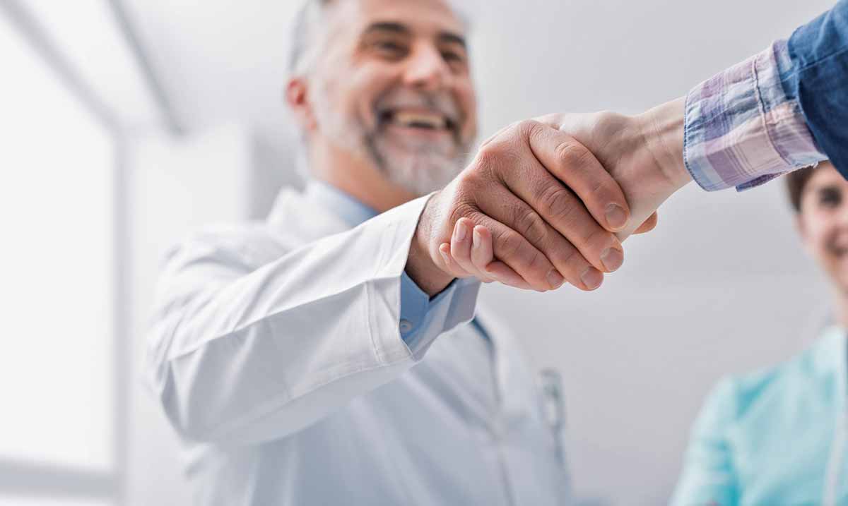 Doctor and female patient meeting at the hospital shake hands. The doctor has a beard and is wearing a white lab coat.