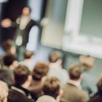 Out of focus image of man standing on a stage and speaking in front of a large audience. Backs of the audience members heads are visible. They are seated in chairs.