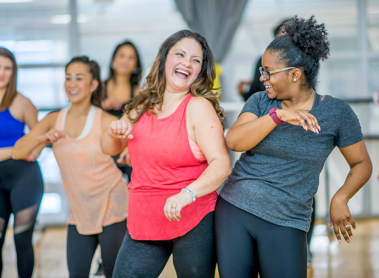 A multi-ethnic group of adult women are dancing in a fitness studio. They are wearing athletic clothes. Two women are laughing while dancing together.