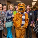 Six women smile with the NIttany Lion mascot at the Lime Spring Outpatient Center. To their right is a table with hors’doeuvres. Behind them is a curtain.