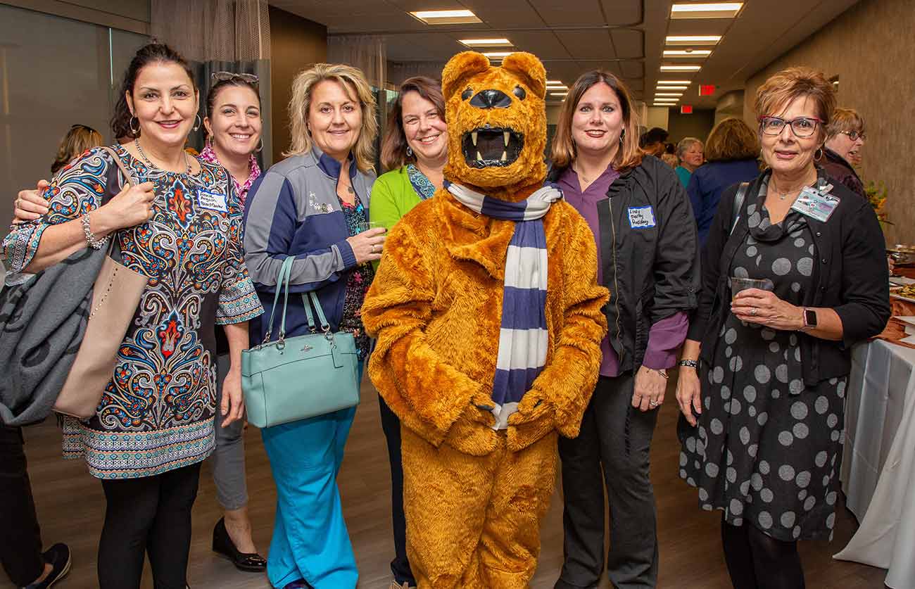 Six women smile with the NIttany Lion mascot at the Lime Spring Outpatient Center. To their right is a table with hors’doeuvres. Behind them is a curtain.