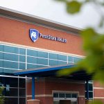 The front entrance of the new Penn State Health Medical Group - Lime Spring Outpatient Center shows a red brick building with the Penn State Health logo, large windows, an entrance roof and tree branches in the foreground.