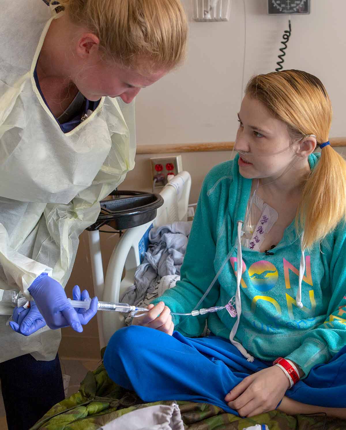 Alyssa Kibler sits in her hospital bed at Penn State Health Milton S. Hershey Medical Center, holding some clear tubing in her hand that is connected to a vial of medicine that registered nurse Amy Koffman has in her hands. Koffman wears a disposable gown and blue gloves as she leans down toward Kibler. Kibler is wearing a sweatshirt and has her hair in a ponytail.