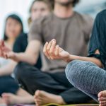 A row of diverse people sit with their legs crossed in a meditation pose. The first woman’s body only is visible. The rest of the people are out of focus. They are wearing yoga pants or sweatpants.