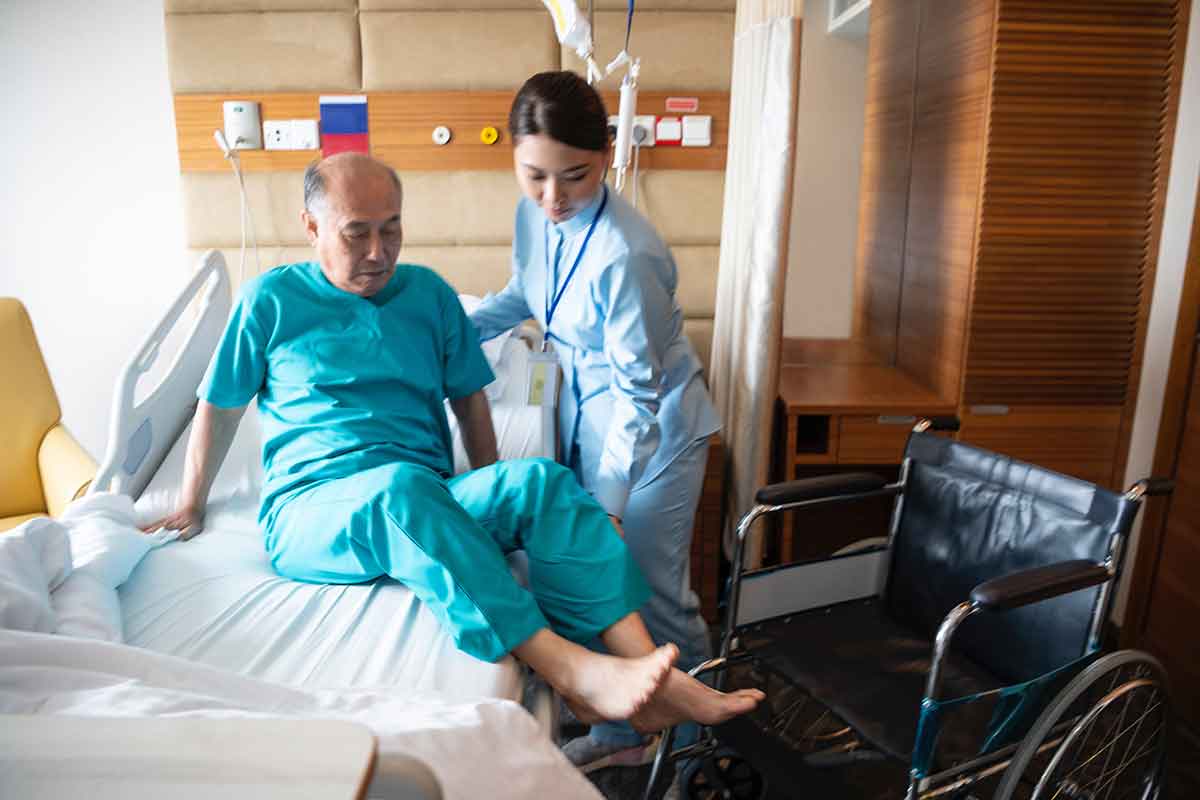 A nurse helps a senior man out of a hospital bed and into a wheelchair. The man is wearing hospital scrubs and has bare feet.