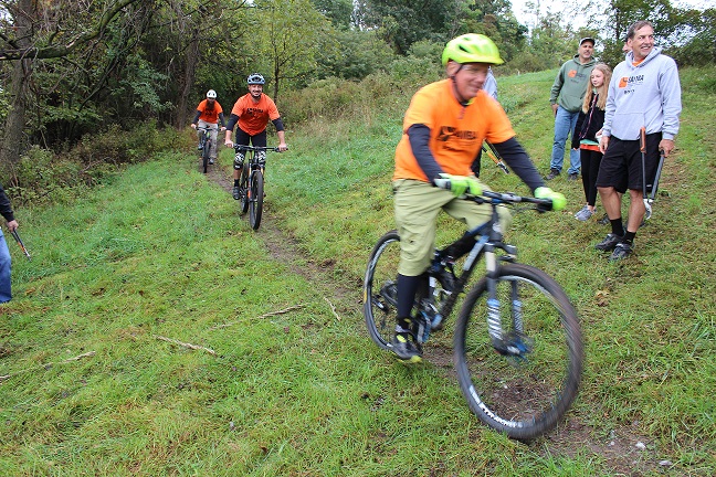 Three men wearing bike helmets and T-shirts ride mountain bikes on a narrow, dirt trail. Four people watch them on either side of the trail. One man is holding garden shears.