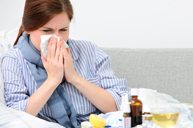 A woman blows her nose with a tissue. In the foreground is a table with various liquid medications and pills, as well as a drink and other items.