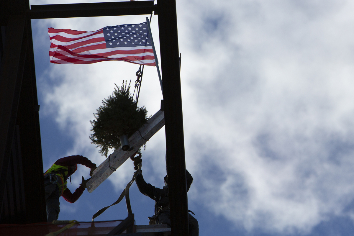 Two construction workers work to secure a white beam, onto which an evergreen tree and American flag are mounted, atop a building.