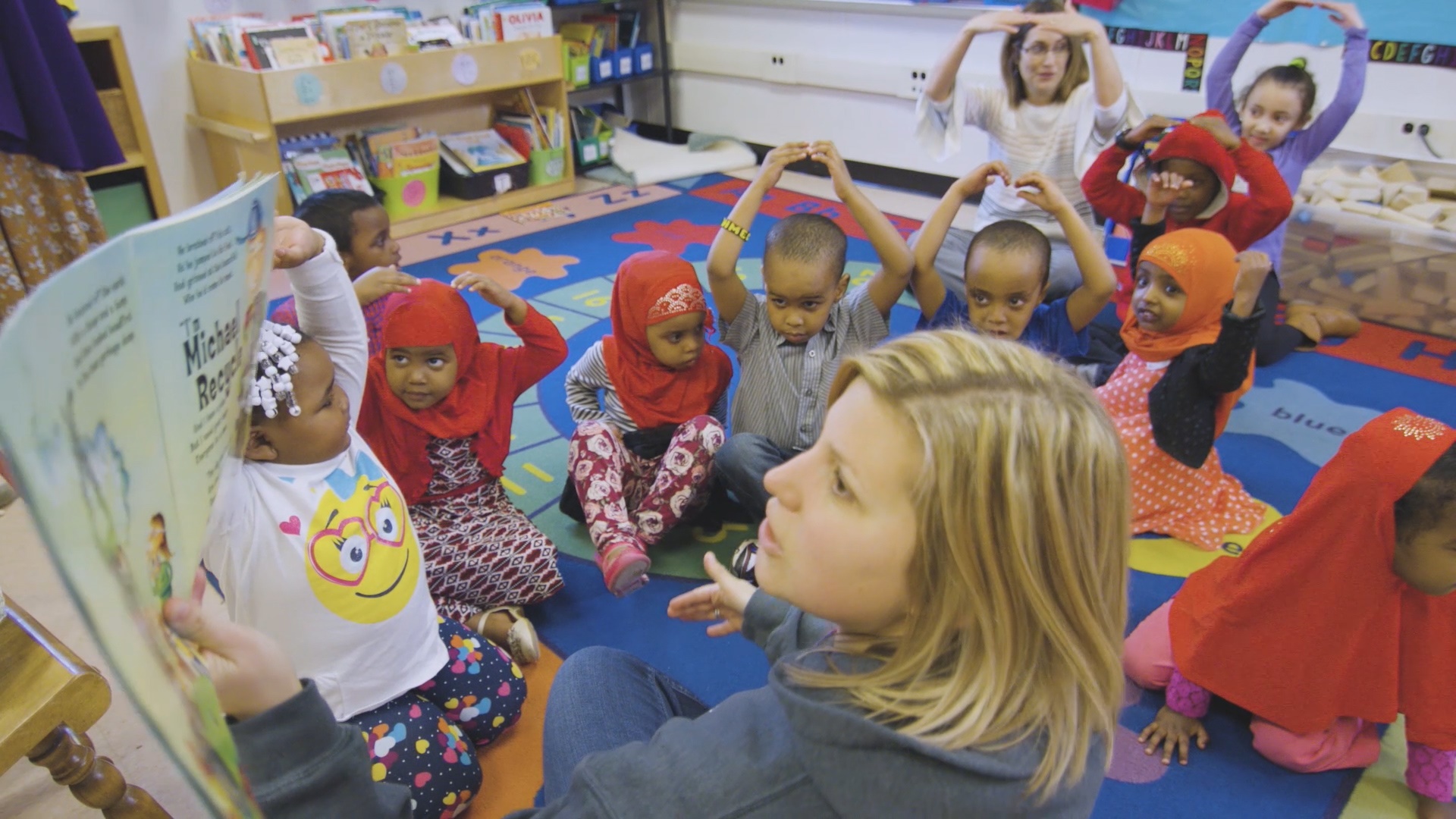 A day care teacher reads a book to 10 diverse preschool children. Another teacher puts her arms above her head with the children. They are all sitting on a colorful rug. Behind them is a bin of books and a bin of wooden blocks.