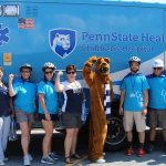 Ten men and women and one boy smile and pose with the Nittany Lion in front of a Penn State Health Children’s Hospital ambulance at the United Way Trike Race. The women and Nittany Lion have their right arms up to make a muscle. One man holds a pom-pom up.