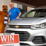 The Nittany Lion mascot and Dr. Craig Hillemeier, dean, CEO and senior vice president for health affairs, give thumbs up next to a 2019 Chevrolet Trax SUV. The Lion is wearing a striped scarf. Dr. Hillemeier is wearing a polo shirt and glasses. In front of the SUV is a sign that says “Win this vehicle.”
