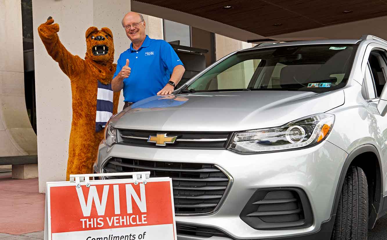 The Nittany Lion mascot and Dr. Craig Hillemeier, dean, CEO and senior vice president for health affairs, give thumbs up next to a 2019 Chevrolet Trax SUV. The Lion is wearing a striped scarf. Dr. Hillemeier is wearing a polo shirt and glasses. In front of the SUV is a sign that says “Win this vehicle.”