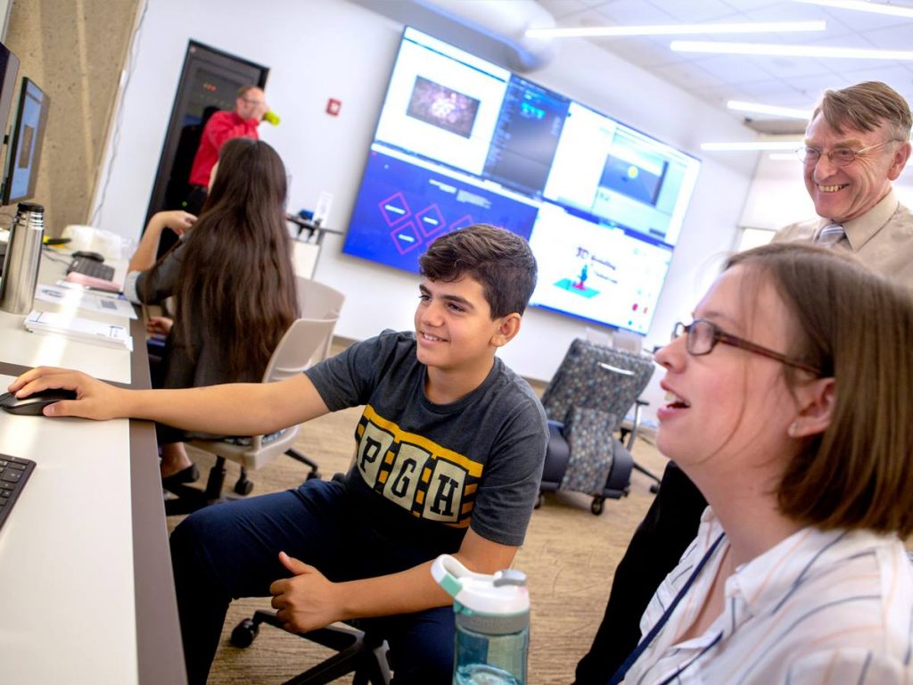 With Seamus Carmichael looking on, rear, 14-year-old Mahmut Esat Yerlikaya gets some hands on experience from Eliza Donne, right, on game creation during the Day of Making event at Harrell Health Sciences Library in August 2018. The students are pictured sitting in front of a computer and laughing, while an adult man looks on.
