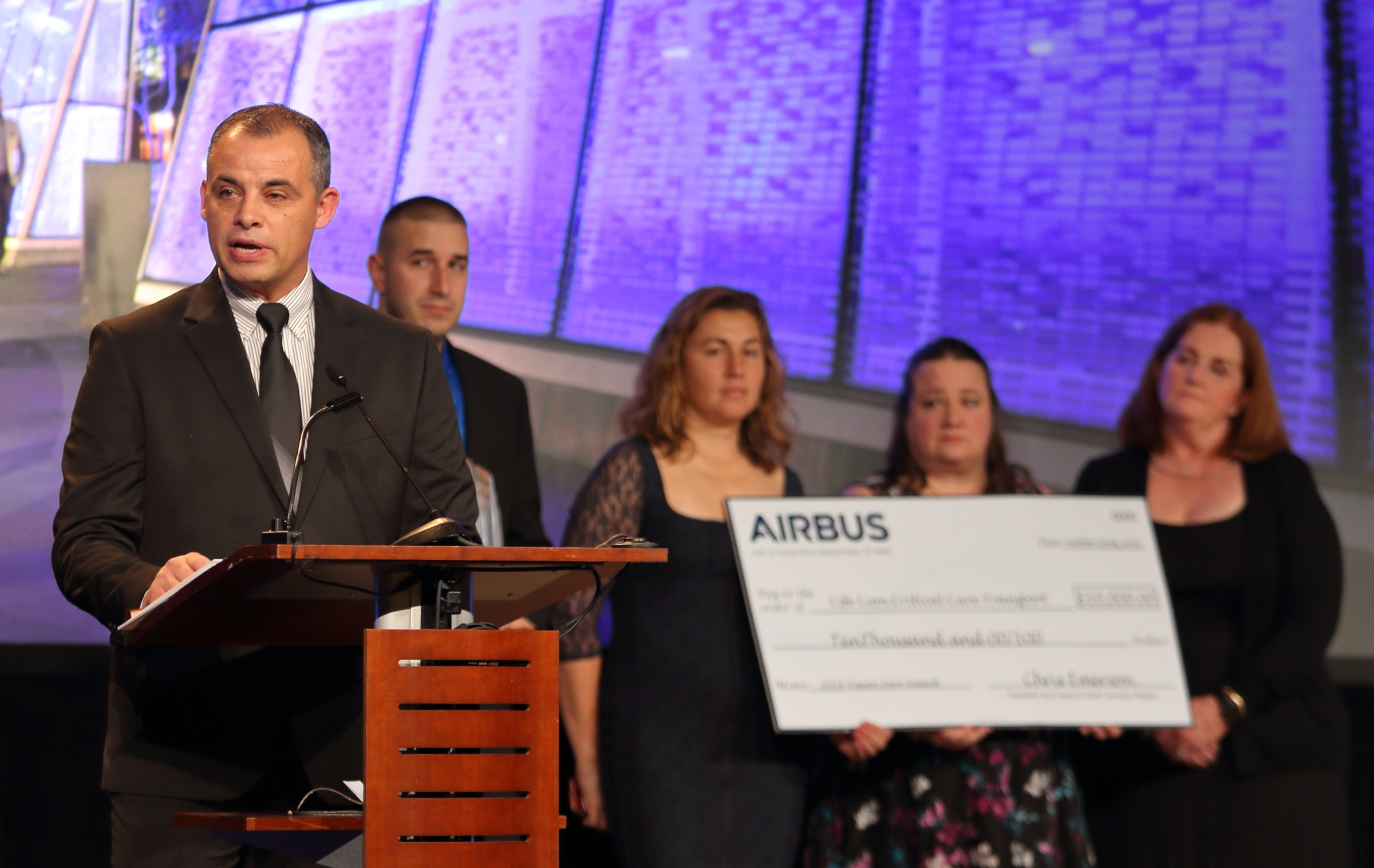 A man in a suit stands at a lectern speaking. Four people stand behind him holding an oversized cardboard check.