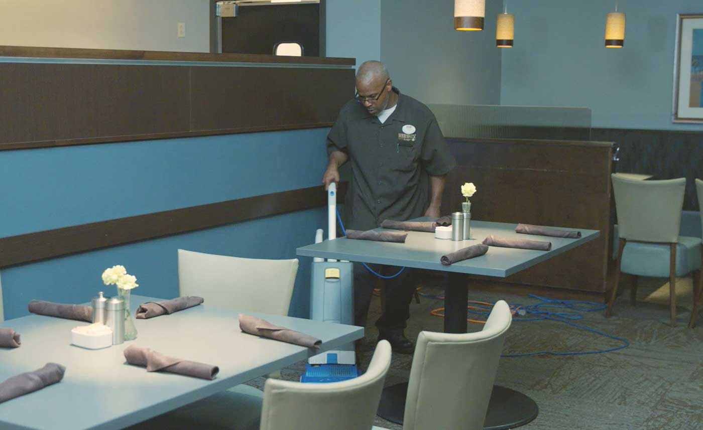 A man vacuums under a table in a restaurant. He is wearing glasses and a shirt with a Hershey Lodge logo and a nametag on it. Two tables with napkins on them are in front of him. A wall with strips of paneling is to his left, and another table is behind him. Lights are visible above him.
