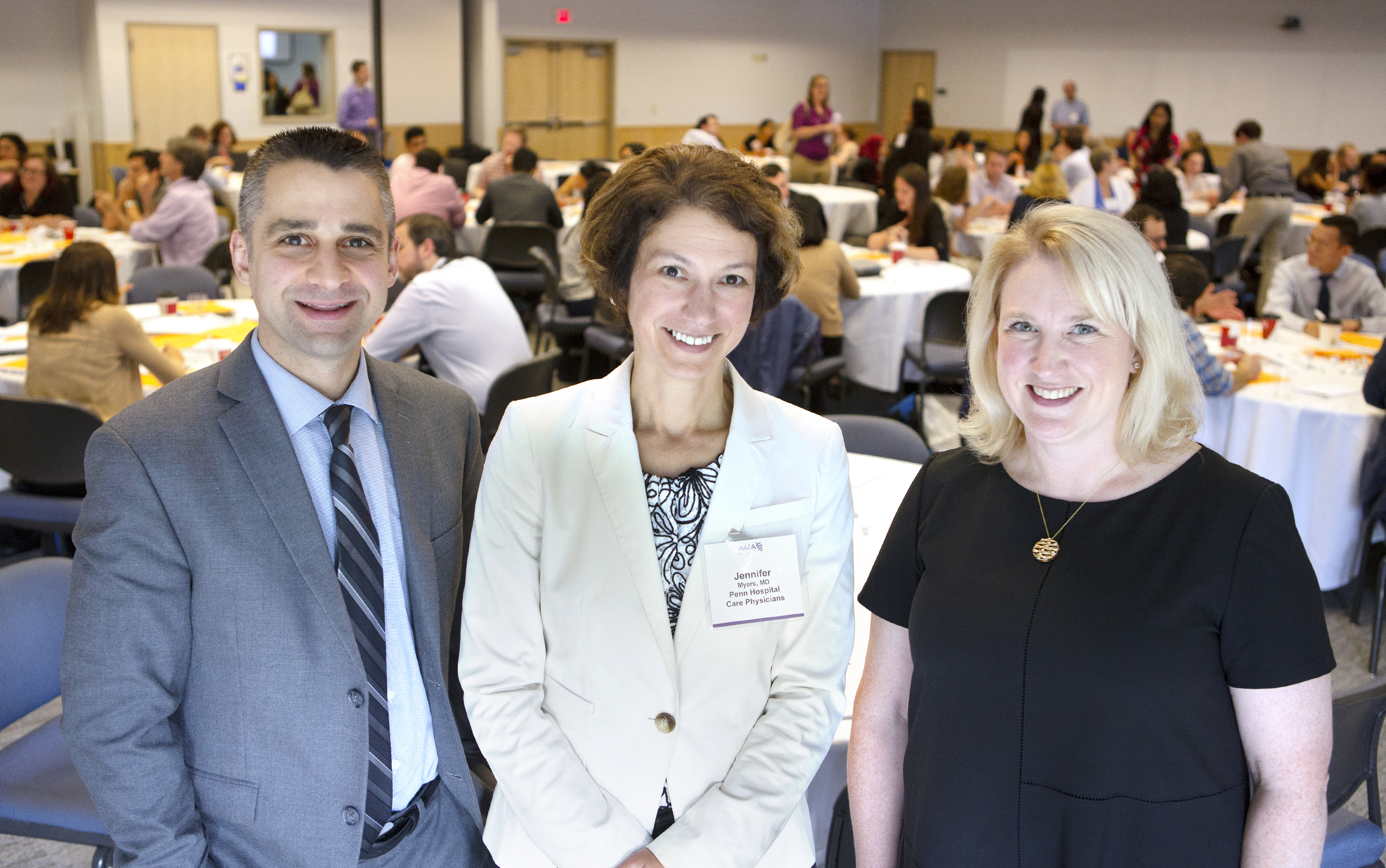 Dr. Jed Gonzalo, Dr. Jennifer Myers and Dr. Luan Lawson, the keynote speakers during the American Medical Association conference, stand and smile. Behind them are people at round tables.