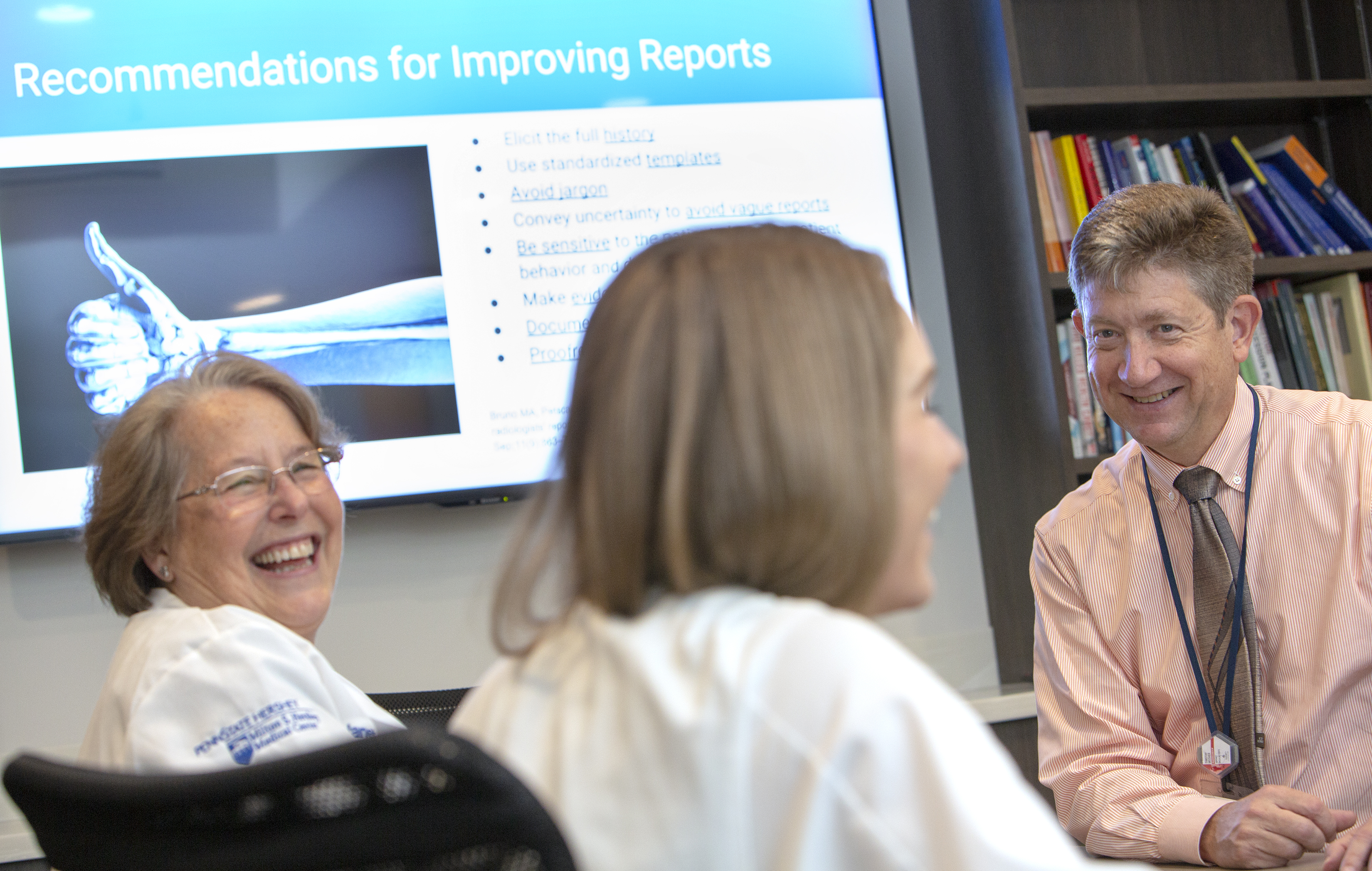 Dr. Timothy Mosher, chair of the Department of Radiology at Penn State College of Medicine, smiles. Two women wearing white lab coats with the Milton S. Hershey Medical logo on them, smile. Behind them is a PowerPoint slide that says “Recommendations for Improving Reports” and has an image of an arm with a thumb up. Behind Dr. Mosher is a bookshelf with books.