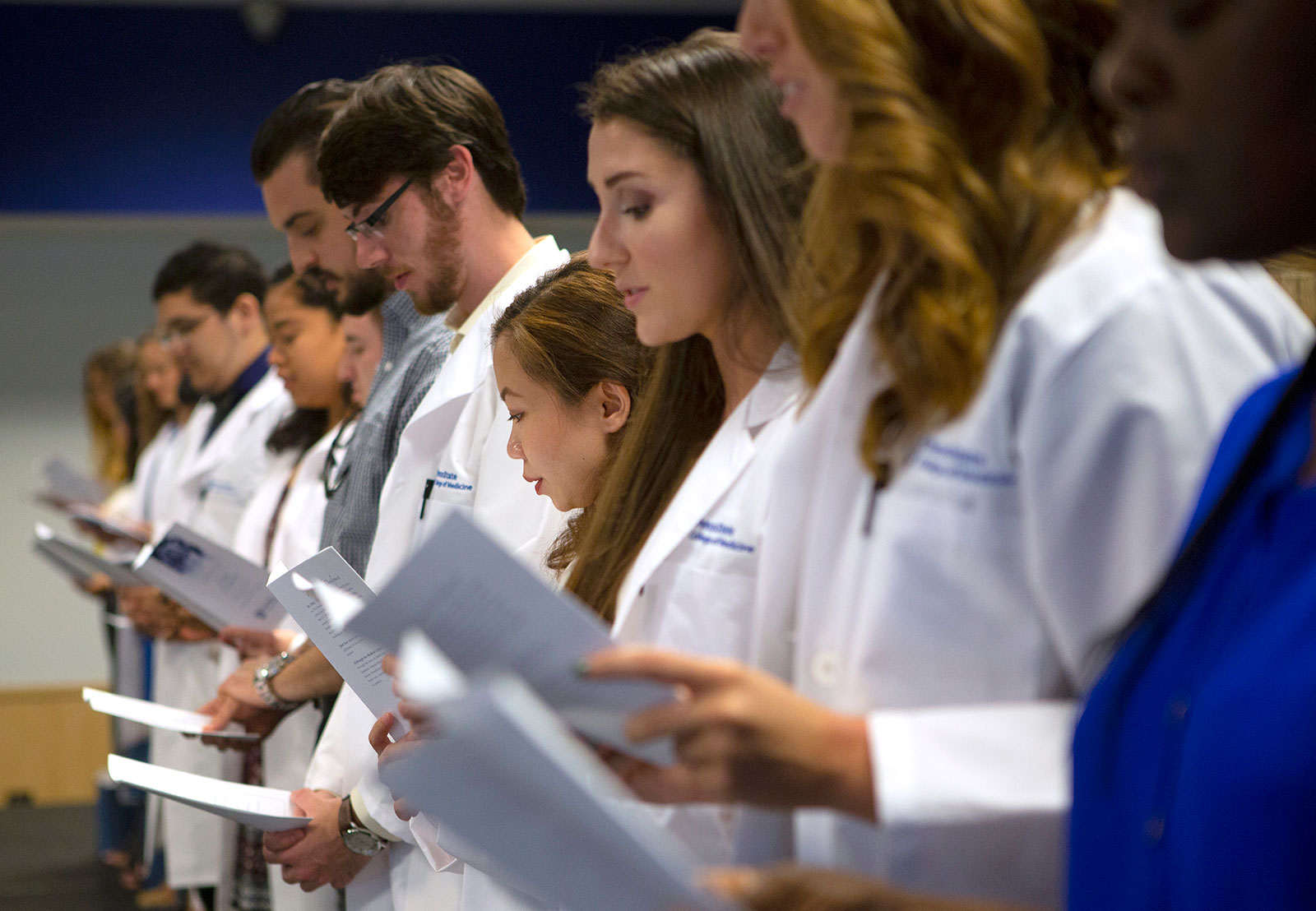 Participants on stage recite the Graduate Student Oath during the Penn State College of Medicine Graduate Oath Ceremony in August 2018. A line of students is pictured holding a paper with the oath printed on it. The students are looking down and reading from the paper.