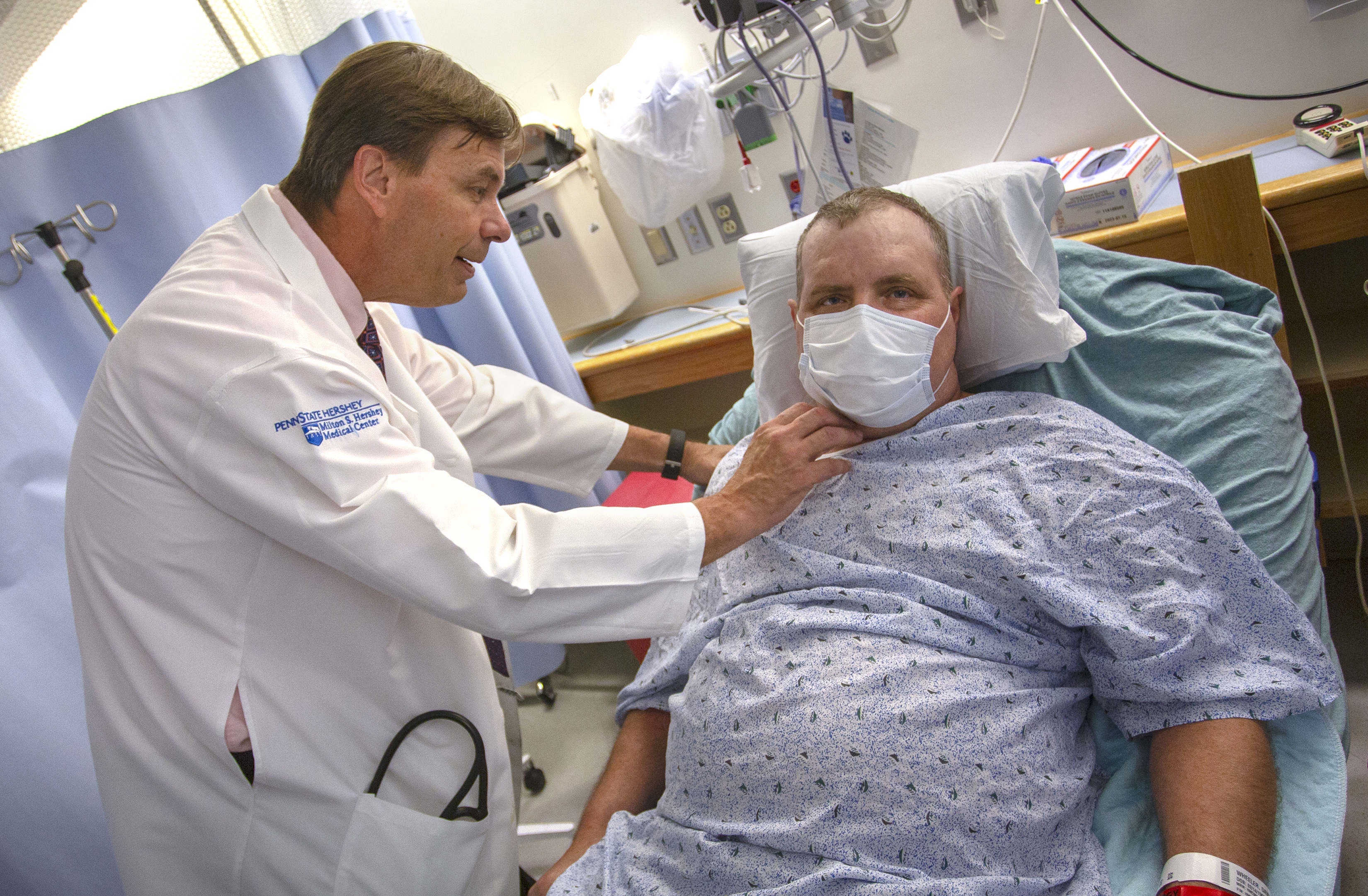 Dr. John Boehmer, left, touches David Wheeler’s neck. Boehmer is wearing a white lab coat with the Hershey Medical Center logo on it. Wheeler is propped up on a hospital bed and is wearing a hospital gown and surgical mask. Behind them is a shelf with medical equipment and cables.