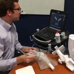 Penn State College of Medicine medical student Jason Spicher is seen using his 3D-printed ultrasound phantom to practice giving ultrasound-guided knee injections. Spicher is pictured sitting at a table with a knee model in front of him, looking at a machine with a keyboard and ultrasound image on it.