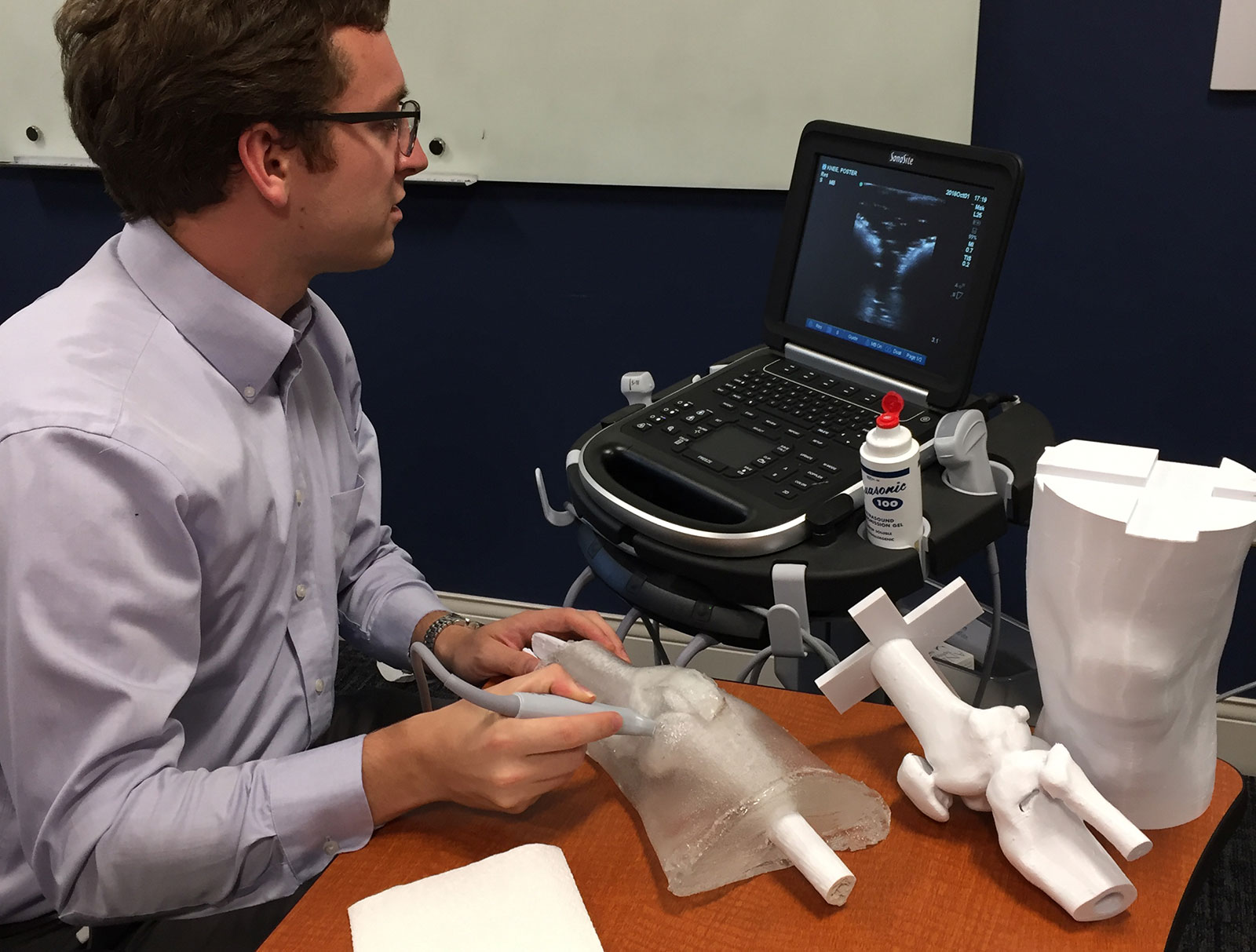 Penn State College of Medicine medical student Jason Spicher is seen using his 3D-printed ultrasound phantom to practice giving ultrasound-guided knee injections. Spicher is pictured sitting at a table with a knee model in front of him, looking at a machine with a keyboard and ultrasound image on it.