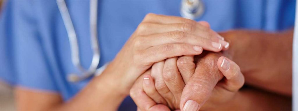 Hands of woman doctor hold the hands of a man. The doctor is visible in the background wearing scrubs and a stethoscope.