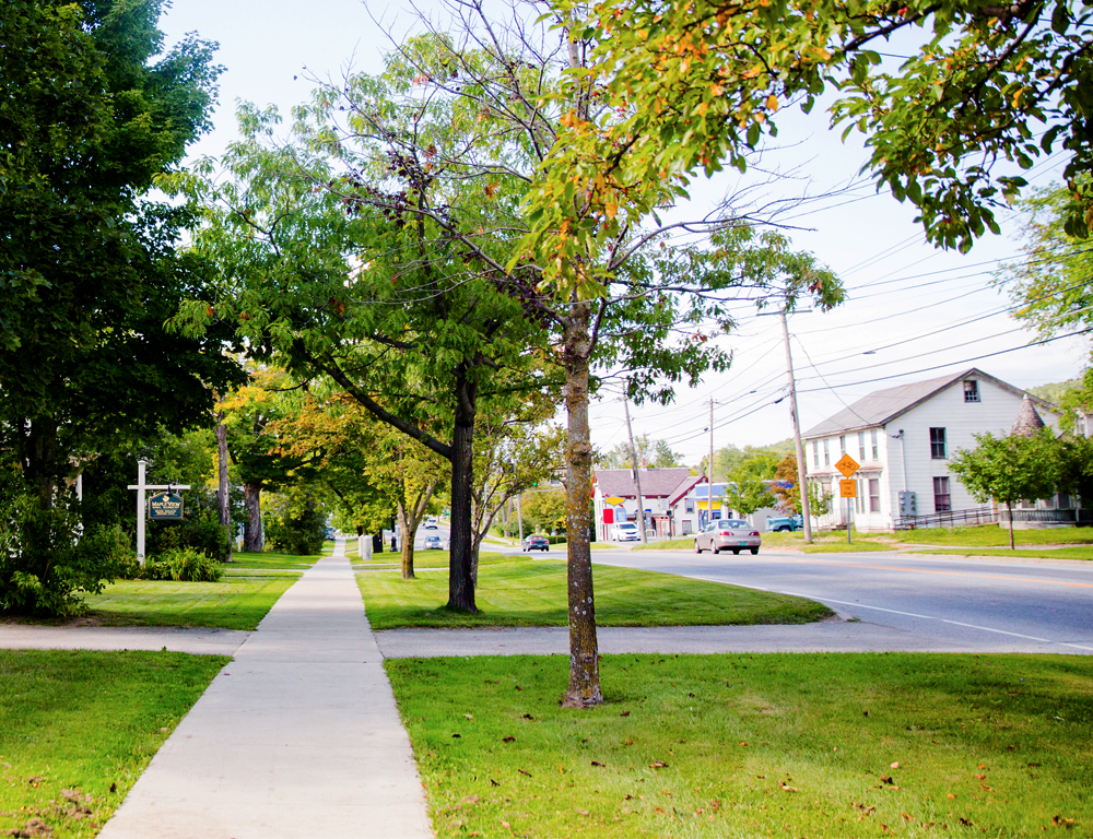 The view down a long sidewalk of a residential neighborhood. A grassy area lined with trees is between the sidewalk and the road. Some cars and buildings are in the background.