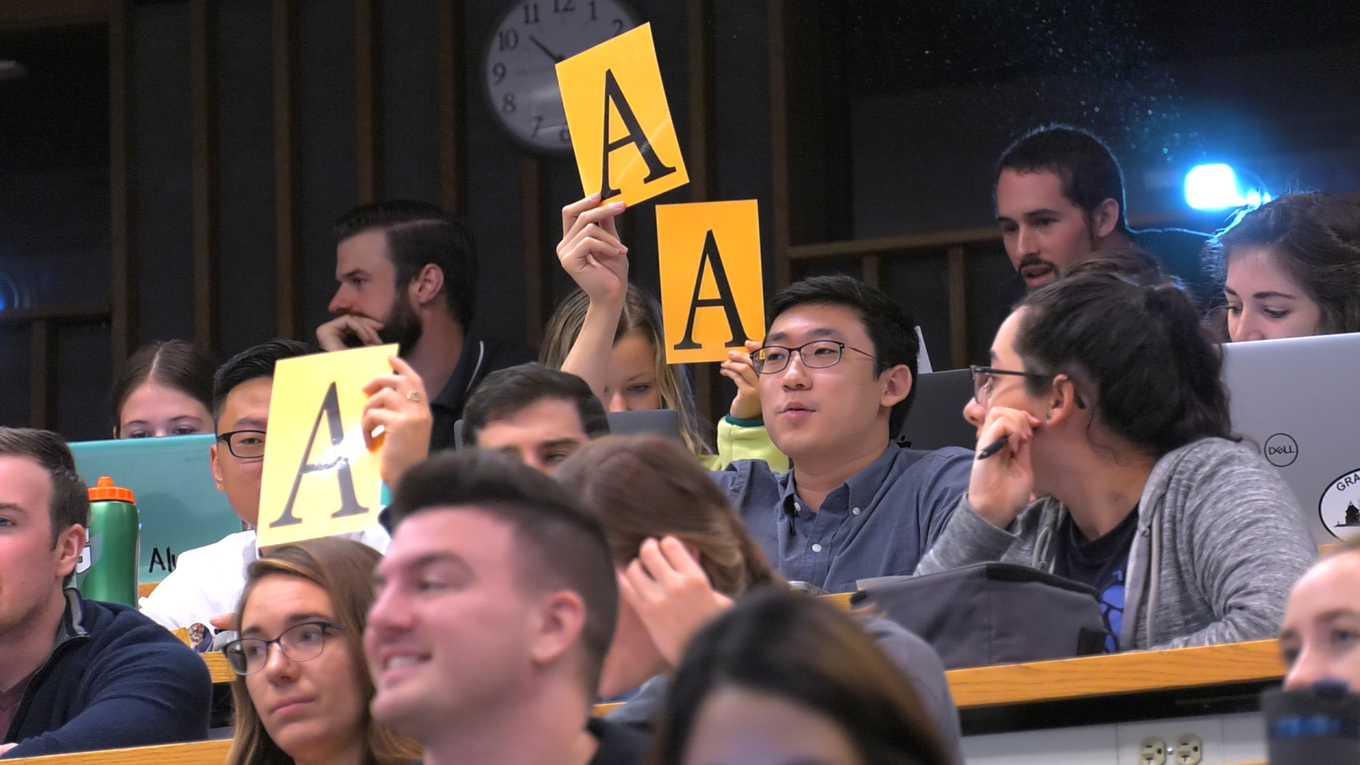 A group of approximately 12 Penn State College of Medicine students sit in a classroom. Two of them hold up the letter “A” on a large piece of paper. Some are smiling. Behind them is a clock and wooden studs on the wall.