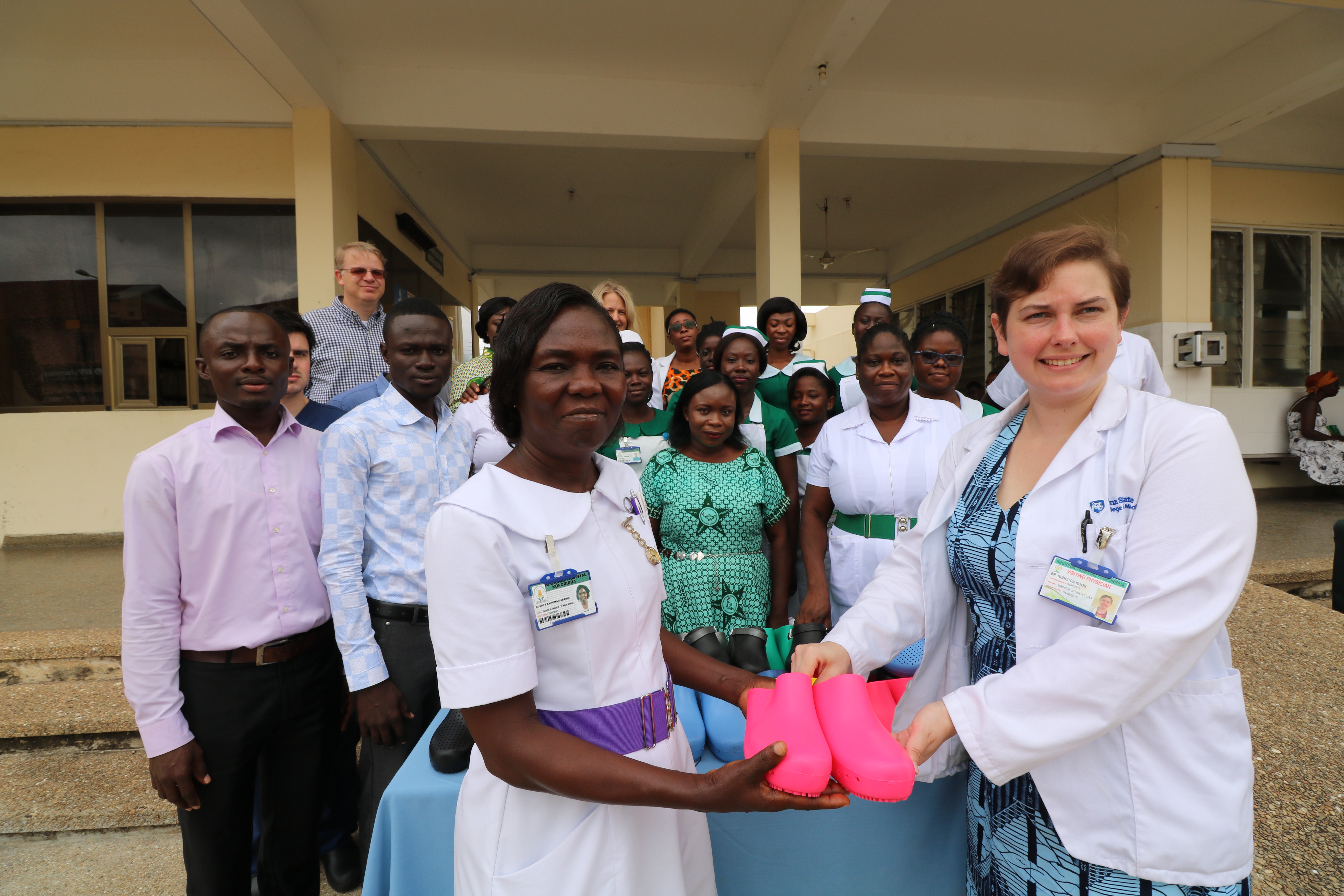 Penn State College of Medicine Global Health Scholars student Becky Koob presents a pair of pink hospital-grade shoes to Gladys Ampomah-Ababio, head of nursing at Eastern Regional Hospital, Ghana, outside the hospital building. Koob wears a white lab coat over her dress, and Ampomah-Ababio wears a white nurse’s uniform with a purple belt. Other hospital staff stand behind a table of donated shoes, smiling. Richard Yeboako, deputy human relations manager and international relations coordinator at the hospital, stands beside the table.