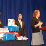 Pediatric Trauma and Injury Prevention manager Amy Bollinger, right, a woman with shoulder-length hair, stands at a podium discussing unsafe toys at a press conference. Behind her stand Reuben Mathew of PennPIRG, left, and Pennsylvania Auditor General Eugene DePasquale, right. Between the two men is a table with wrapped gifts.