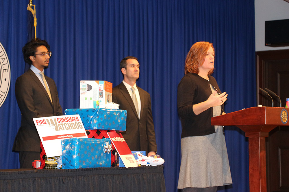 Pediatric Trauma and Injury Prevention manager Amy Bollinger, right, a woman with shoulder-length hair, stands at a podium discussing unsafe toys at a press conference. Behind her stand Reuben Mathew of PennPIRG, left, and Pennsylvania Auditor General Eugene DePasquale, right. Between the two men is a table with wrapped gifts.