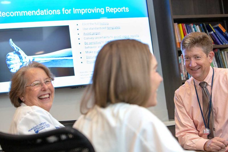 Dr. Timothy Mosher, chair of the Department of Radiology at Penn State College of Medicine, smiles. Two women wearing white lab coats with the Milton S. Hershey Medical logo on them, smile. Behind them is a PowerPoint slide that says “Recommendations for Improving Reports” and has an image of an arm with a thumb up. Behind Dr. Mosher is a bookshelf with books.