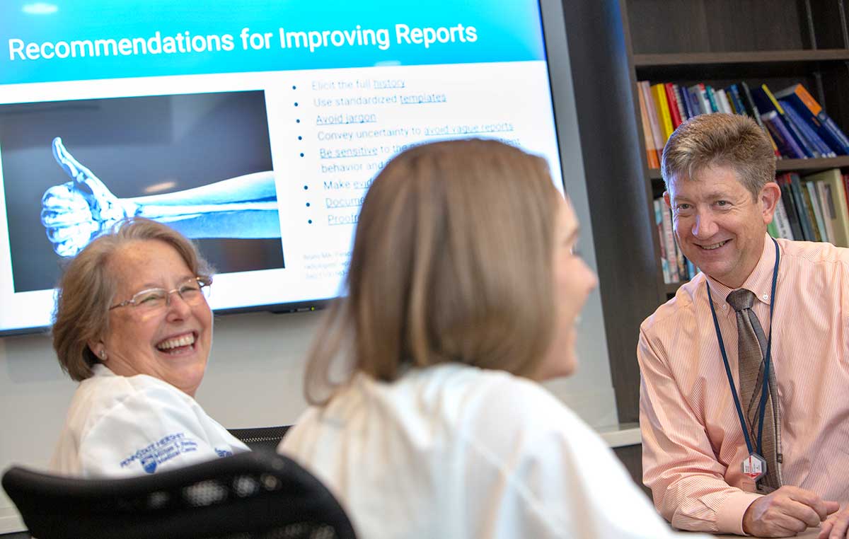 Dr. Timothy Mosher, chair of the Department of Radiology at Penn State College of Medicine, smiles. Two women wearing white lab coats with the Milton S. Hershey Medical logo on them, smile. Behind them is a PowerPoint slide that says “Recommendations for Improving Reports” and has an image of an arm with a thumb up. Behind Dr. Mosher is a bookshelf with books.