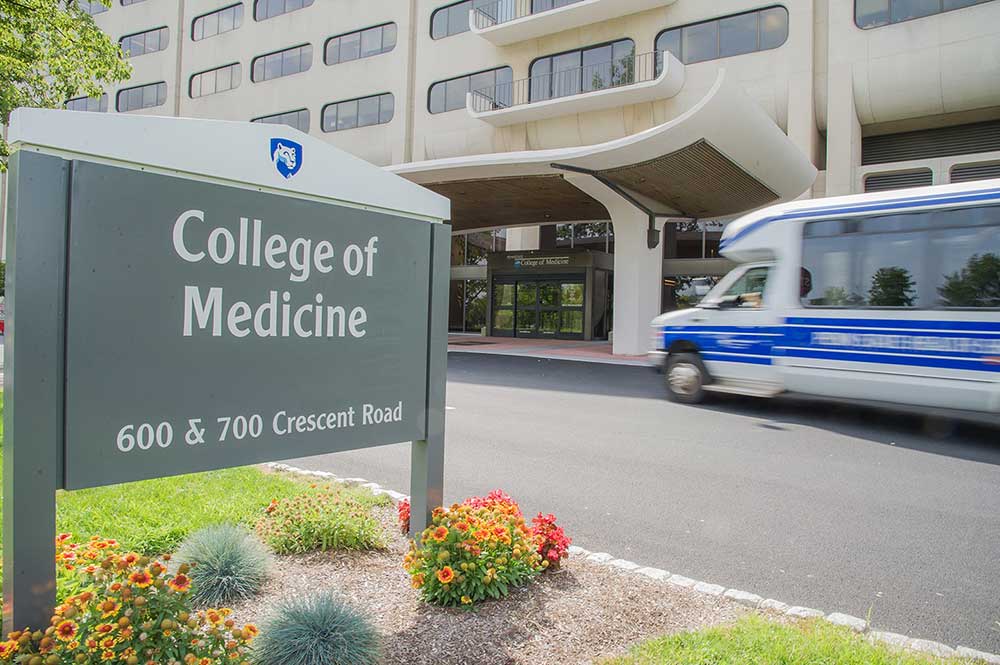 A shuttle bus drives toward the Penn State College of Medicine entrance. A sign says “College of Medicine, 600 & 700 Crescent Road.” Flowers are below the sign. The college building features rows of long windows.