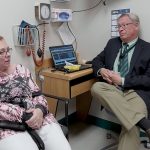 Denise Brown, who suffers from aortic stenosis and needed heart valve replacement surgery, sits with Dr. John Conte at Penn State Milton S. Hershey Medical Center. Brown is sitting in an exam room on a chair next to Conte, who is sitting on a stool next to a desk on which his laptop sits, open. Brown wears a cannula in her nose to assist with breathing.